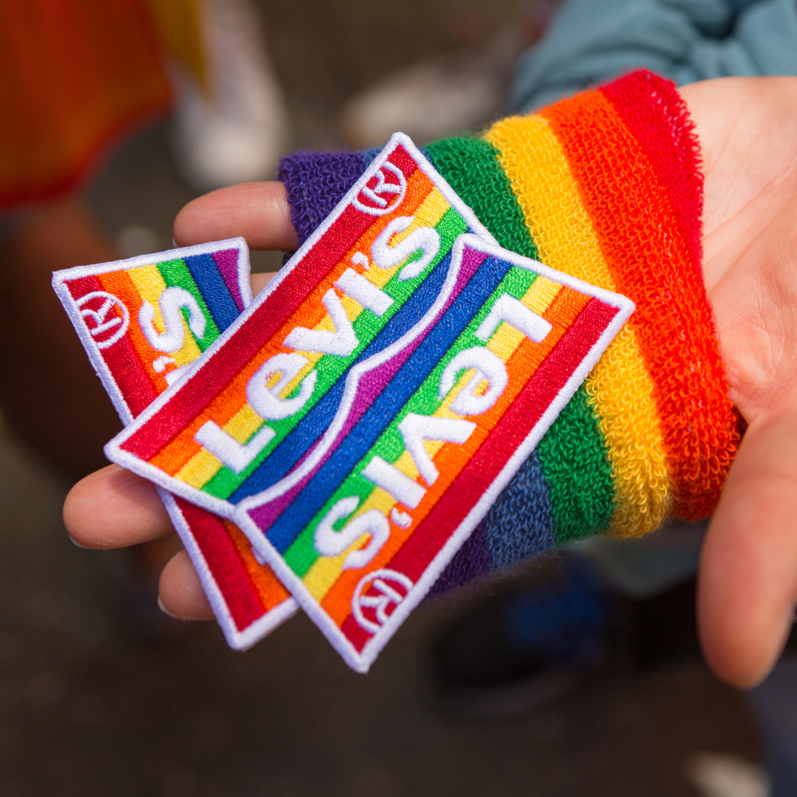 A hand wearing a rainbow fingerless glove holds a handful of Levi's® Pride patches - Rainbow striped patches that are in the shape of the Levi's® batwing logo and read 