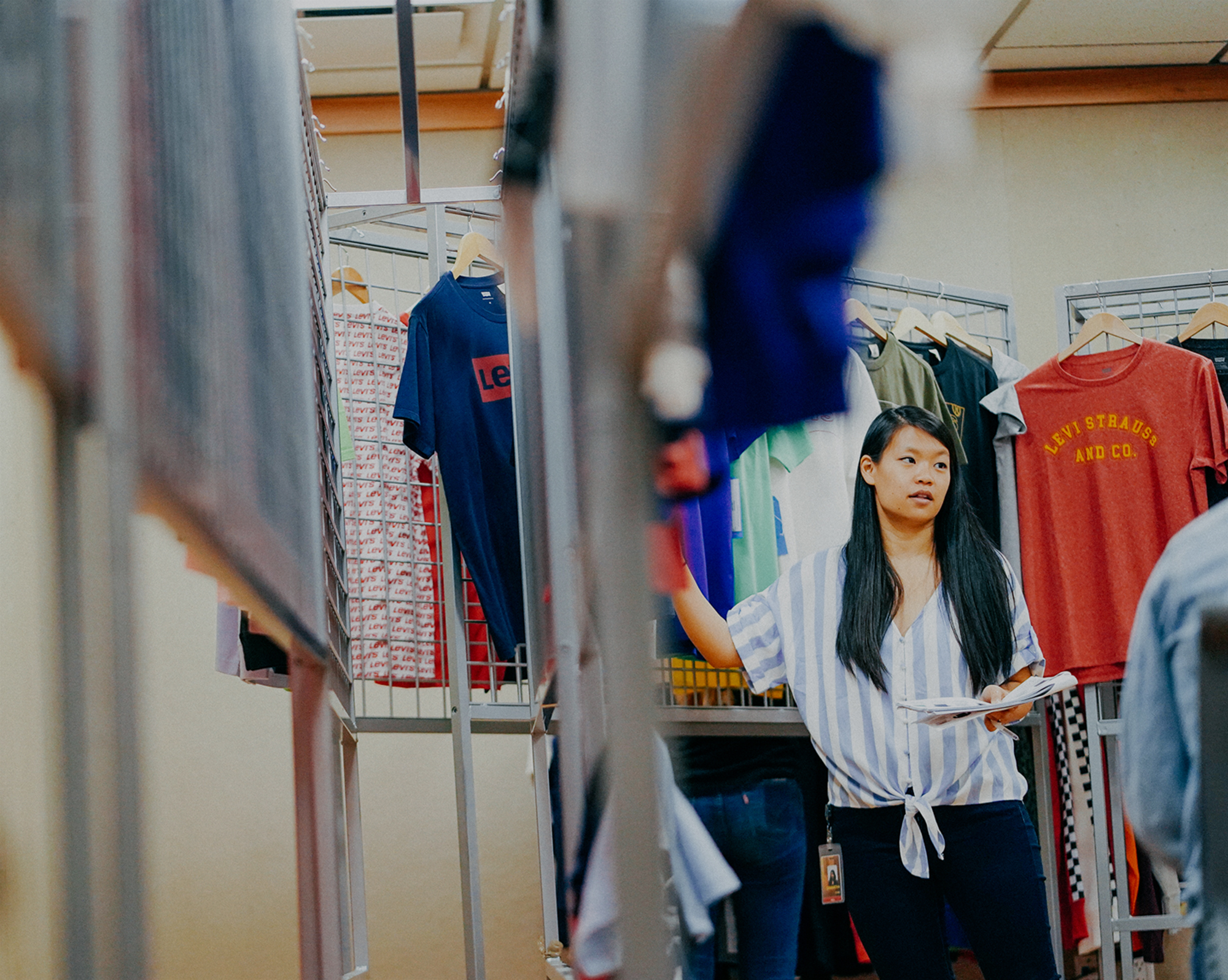 A person with long black hair and a blue and white vertical striped shirt stands next to clothing racks of Levi's® products. A blurred clothing rack is in the foreground.