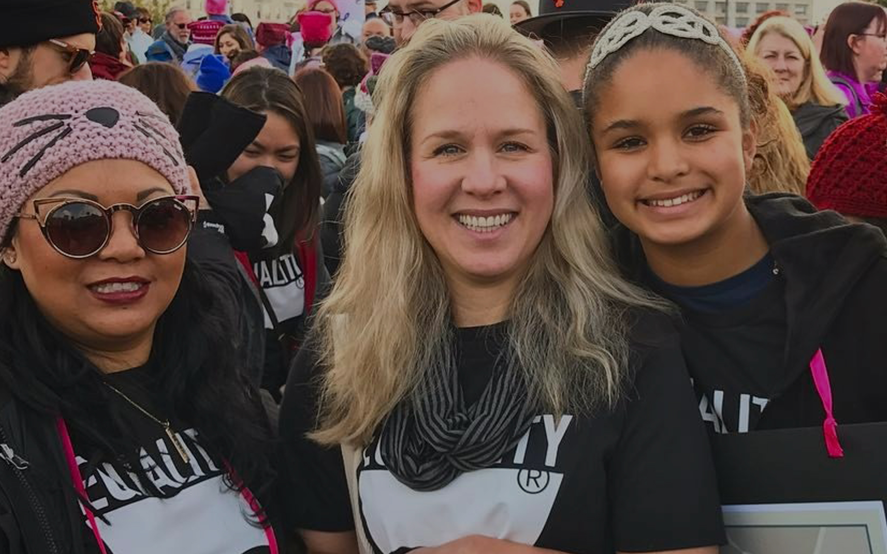 A close-up photo of three smiling women wearing matching black shirts. There is a crowd of people behind them.