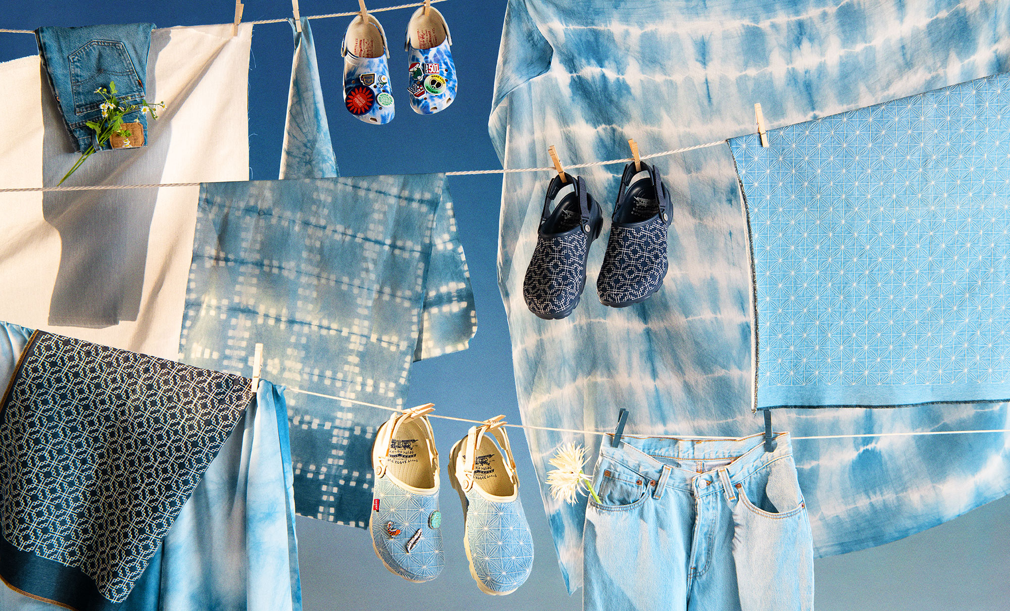 Three pairs of Levi's® Crocs hang on a stacked clothesline next to hanging tie-dye printed fabric.