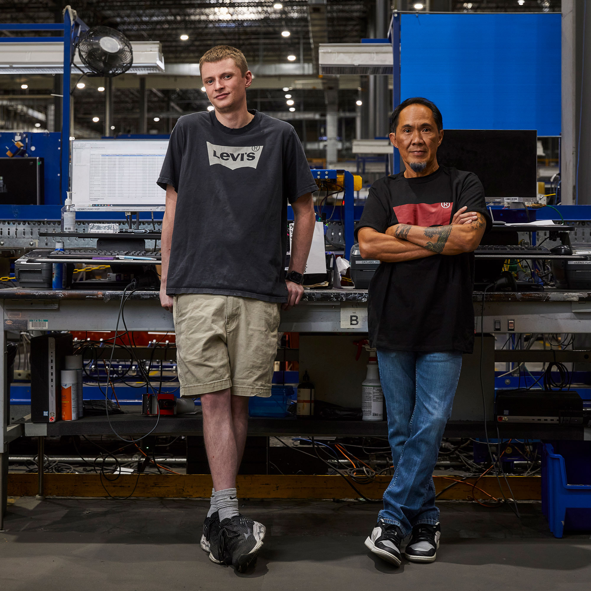 Two people lean on machinery. Person on the left wears a dark grey Levi's® T-shirt featuring a Levi's® batwing logo, and khaki shorts. Their arms are down and hands lean against the machinery. Person on the right wears a dark grey T-shirt with a red Levi's® logo and blue denim jeans. Their arms are crossed in front of their chest.