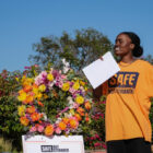 A person stands in holding a piece of paper in a yellow T-shirt that reads "Safe Not Stranded." They are next to a floral wreath that has a sign underneath with the same words.