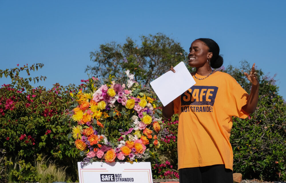 A person stands in holding a piece of paper in a yellow T-shirt that reads "Safe Not Stranded." They are next to a floral wreath that has a sign underneath with the same words.