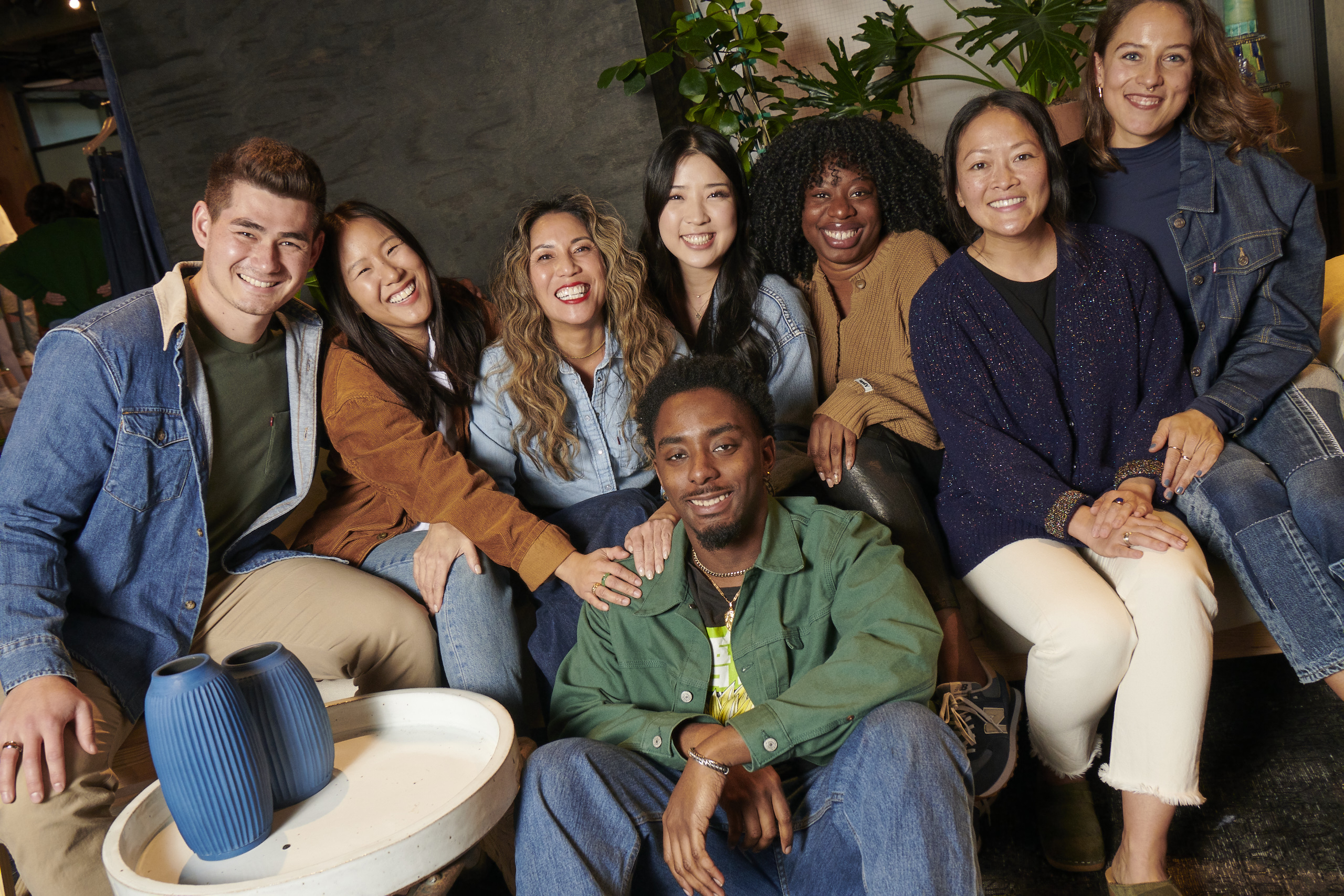 A group of LS&Co. employees sit on a couch and smile. Left to right: a young man with brown hair and a Levi's® sherpa trucker jacket; a young woman with long straight black hair in a tan jacket; a young woman in a chambray button-up with long wavy light brown hair; a young woman with long straight black hair in a denim jacket; a young woman with curly black hair in a tan button up sweater; a young woman with black hair tied back in a sparkly navy sweater and white pants; a young woman with short brown hair in a navy shirt with a denim jacket. A young man with short black hair and a green jacket and blue jeans sits in front of the group.