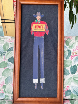 A Levi's® "Shane" cowboy is painted on a denim texture and in a brown rectangular frame. He wears a brown cowboy hat, green neck scarf, a long sleeve red button down and long blue Levi's® jeans. He holds a yellow sign that reads "Levi's America's Finest Overalls." The painting was recently found in a Florida thrift store.