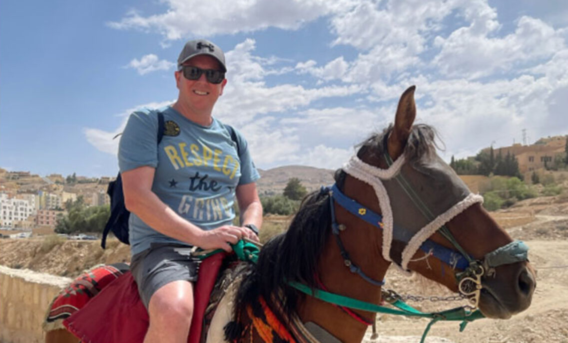 LS&Co. SVP of Global Distribution and Logistics Craig Jones poses on a horse. Craig wears a grey baseball cap, blue T-shirt that reads "Respect the Grind" and grey shorts. The horse is brown, wears blinders and is decorated in multi-color tassels.