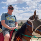 LS&Co. SVP of Global Distribution and Logistics Craig Jones poses on a horse. Craig wears a grey baseball cap, blue T-shirt that reads "Respect the Grind" and grey shorts. The horse is brown, wears blinders and is decorated in multi-color tassels.