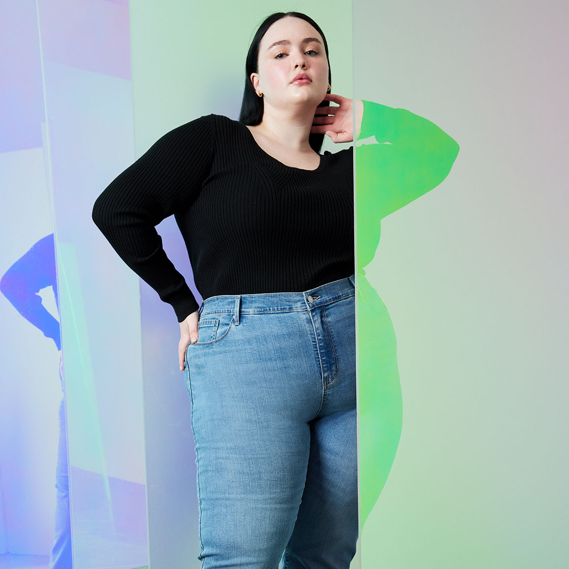 A person with long dark hair faces the camera. Their left arm is bent and their hand is behind their neck. Their right arm is bent and their hand is placed on their hip. They are wearing a black long sleeve and Denizen by Levi Strauss & Co. medium wash jeans. The photo is cropped below their knees. There is an abstract purple and green light-streaked background. Photo by Julia Johnson.