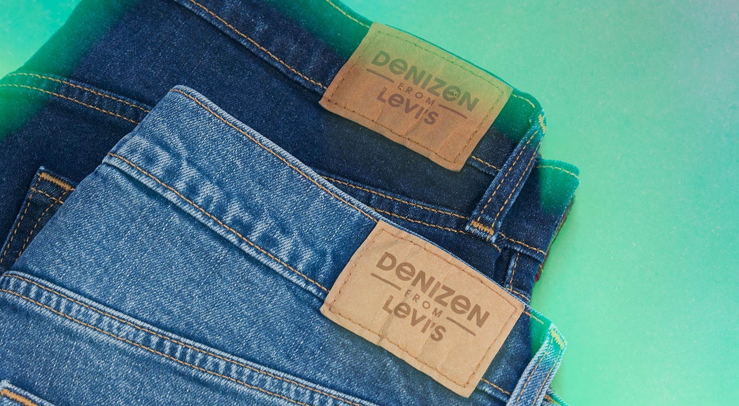 A close up of two folded pairs of Denizen From Levi's® jeans. The one on top is medium wash and the one in the back is dark wash. They are against a green background and both have brown tags on the waistband that read 