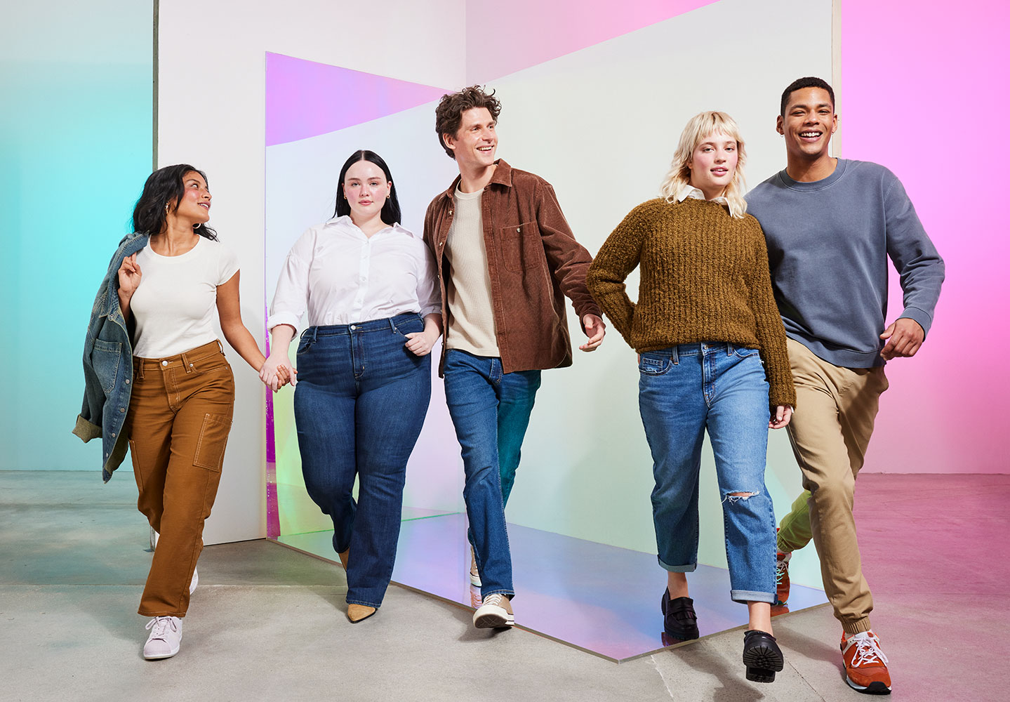 A group of people hold hands against a fluorescent colorful background. They wear Denizen by Levi Strauss products. Person on the left wears a white t-shirt, brown pants and sneakers. They have a blue denim jacket held over their shoulder. The next person wears a long sleeved white button-down tucked into blue jeans. The person in the middle wears a brown button up jacket over a white t-shirt with blue denim jeans. The next person has a white collar poking out of a brown sweater tucked into blue jeans with a rip on the right knee. They wear black loafer shoes. The person on the far right is wearing a long sleeve blue shirt and khaki pants with red sneakers. Photo by Julia Johnson.