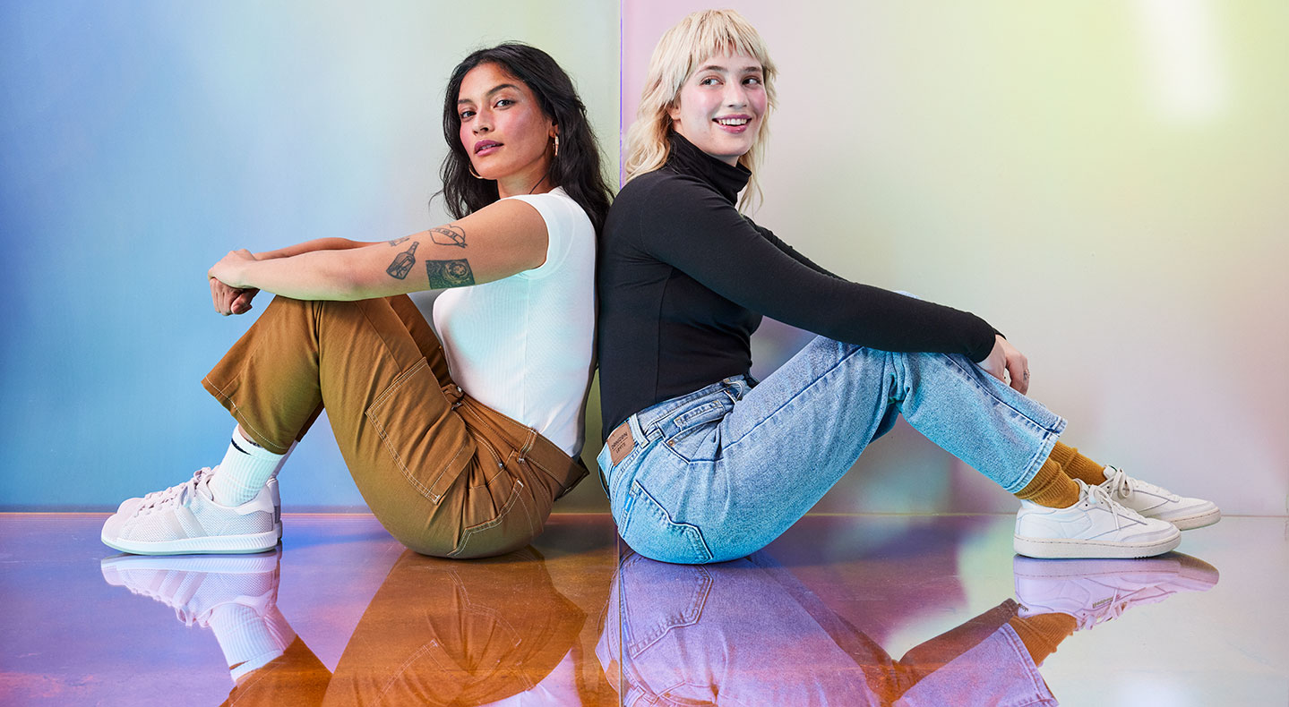 Two people sit on a reflective floor back to back. The person on the right has long dark hair and is wearing a white t-shirt. They have some tattoos on their left arm and are wearing cargo-style tan pants with white socks and sneakers. The person on the right has medium blonde hair and bangs. They are wearing a black turtleneck long sleeve shirt and blue jeans with tan socks and white sneakers. Photo by Julia Johnson.