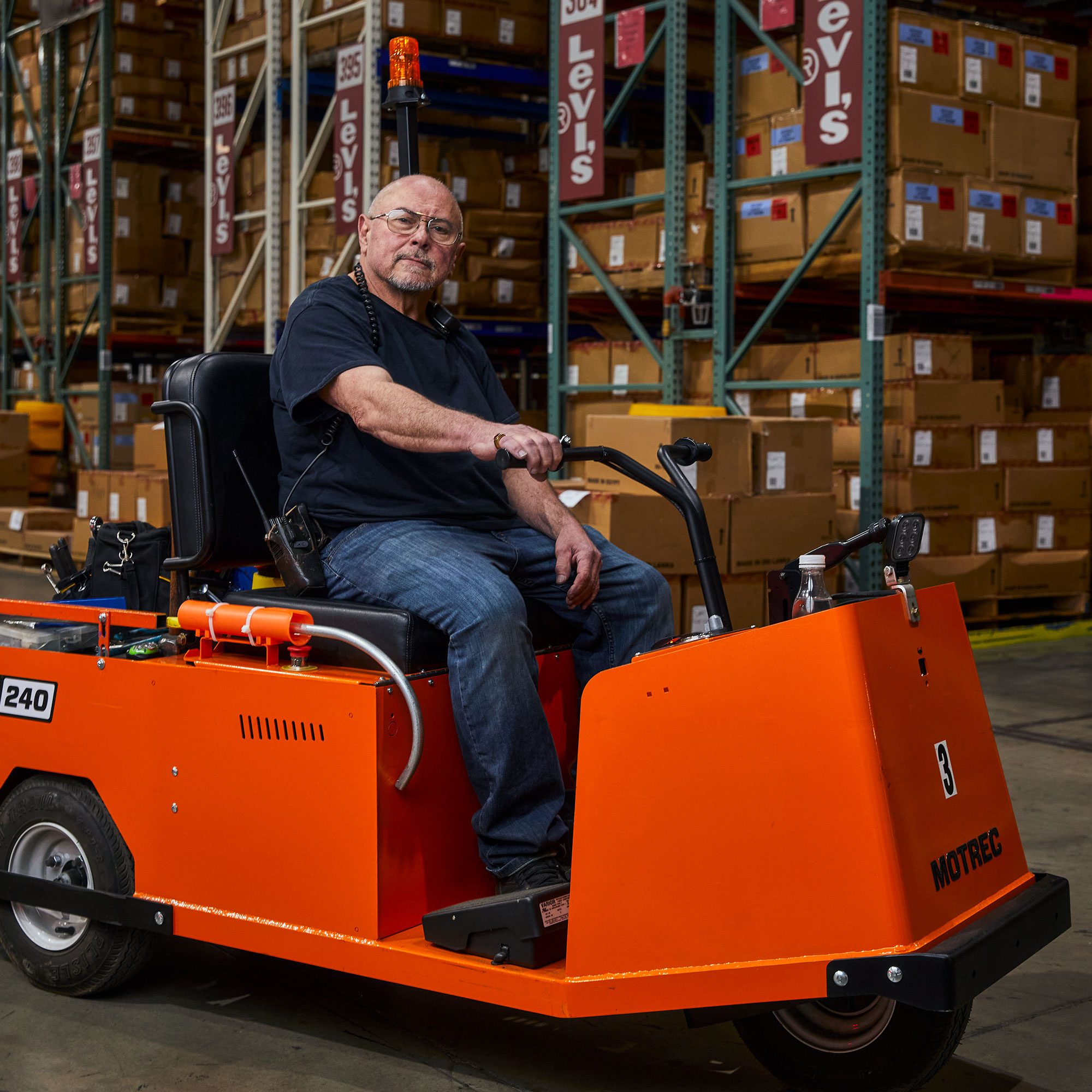 A person sits on an orange distribution center vehicle in a Levi's® distribution center. They are wearing a dark navy T-shirt and blue jeans. There are boxes stacked in the background with Levi's® signs.