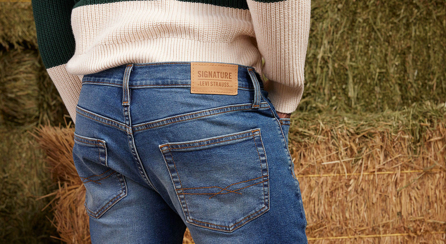 A close up of the back of a person wearing Signature by Levi Strauss jeans. There is a leather patch above the right back pocket with the brand's logo. The person is wearing a knit white sweater and they are facing a hay bale.