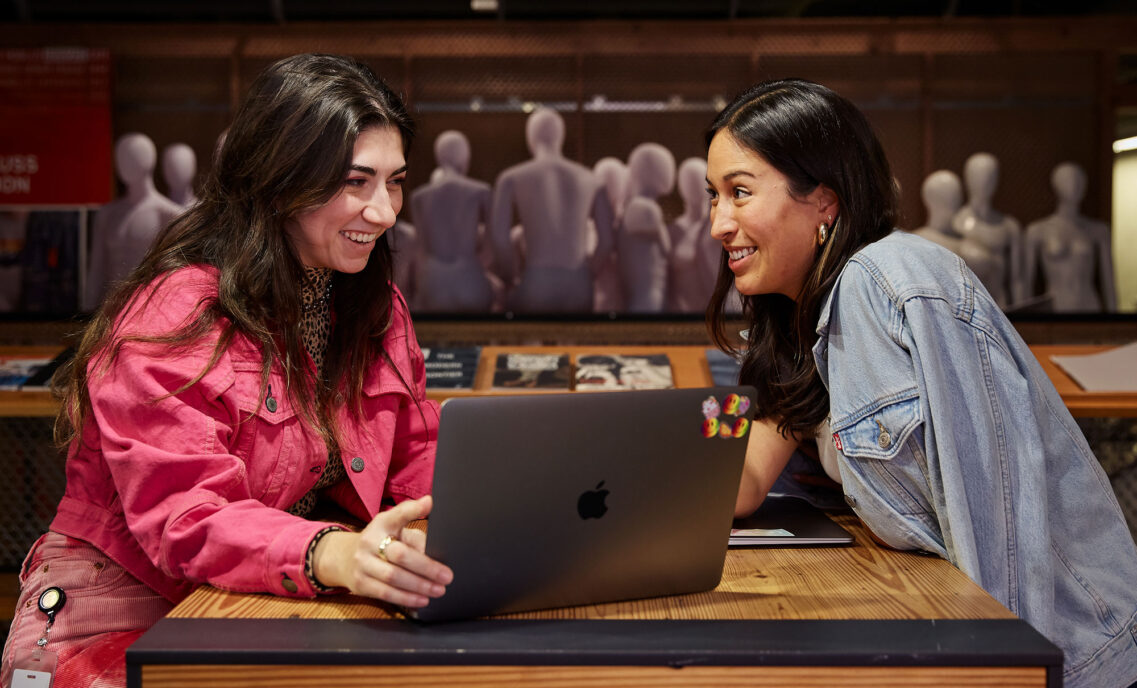 Two LS&Co. employees sit at a table with a laptop in between them. The person on the right is wearing a pink Levi's® jacket and the person on the right is wearing a denim jacket.