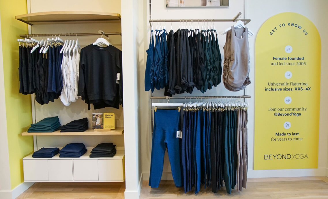 An assortment of athleisure products hang on clothes racks and are stacked in the interior of a Beyond Yoga® store. A yellow sign hangs on the right reading "Get to Know Us. Female founded and led since 2005. Universally flattering, inclusive sizes: XXS-4X. Join our community @BeyondYoga. Made to last for years to come"