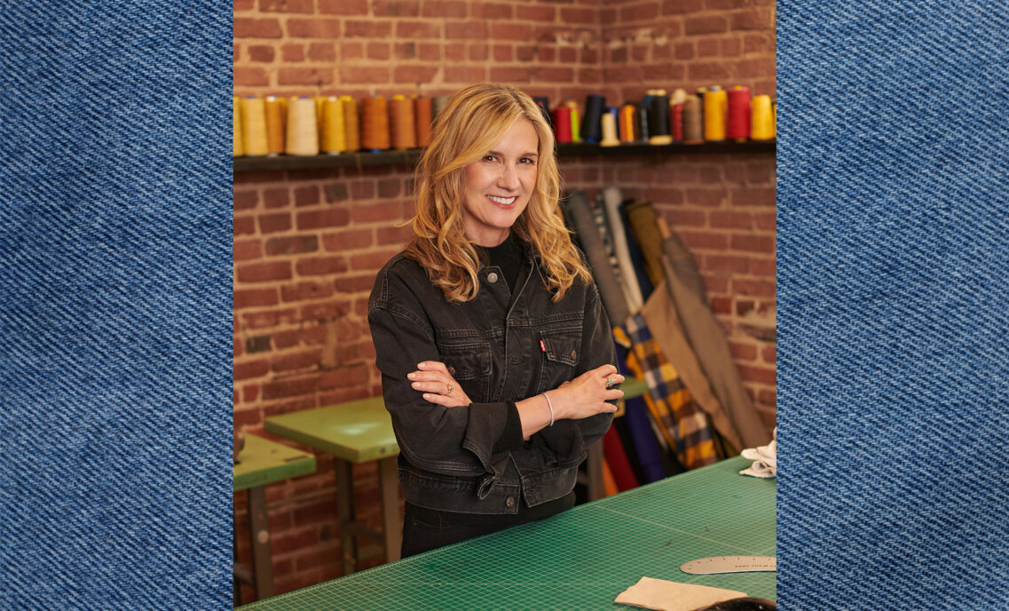 LS&Co. CEO Michelle Gass smiles with her arms crossed. She is wearing a black denim Levi's® jacket. There is a brick wall with spools of thread behind her. Her photo is overlayed on top of a blue denim texture background.