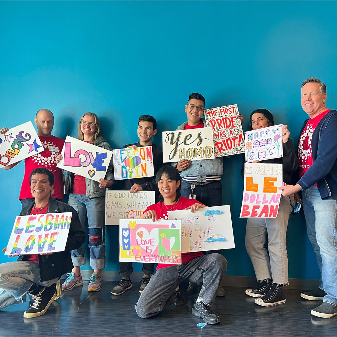 A group of LS&Co. employee volunteers pose with homemade signs at the SF LGBT center featuring words and phrases in support of LGBTQ+ rights.