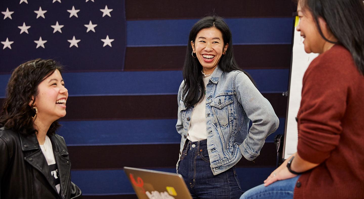 Three employees from the LS&Co. Asian employee resource group (ERG) smile during a meeting in front of a blue and black striped American flag background.