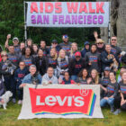 A large group of LS&Co. employee volunteers pose in matching Aids Walk 2019 shirts in front of a sign reading "Aids Walk San Francisco." In front of them is a large sign of the Levi's® pride logo with rainbow coloring.