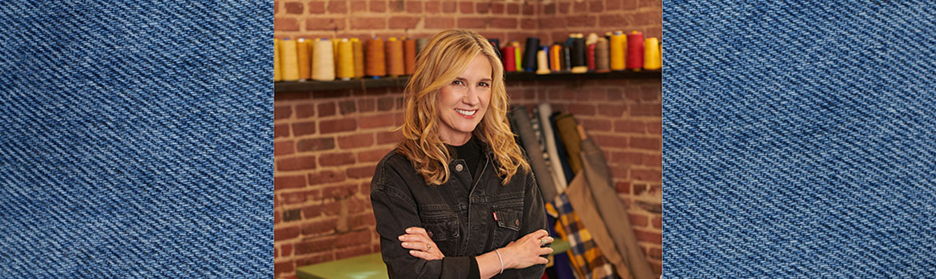 LS&Co. CEO Michelle Gass smiles with her arms crossed. She is wearing a black denim Levi's® jacket. There is a brick wall with spools of thread behind her. Her photo is overlayed on top of a blue denim texture background.