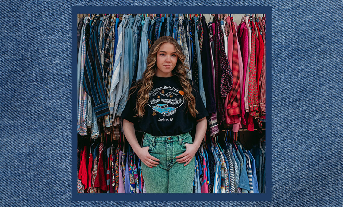 Denim hunter Rose DeBruin stands in front of racks of clothing in her vintage shop. She is smiling and her hands are looped in her front belt loops. She wears a black T-shirt and green pants. The photo is placed over a blue denim texture background.