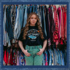 Denim hunter Rose DeBruin stands in front of racks of clothing in her vintage shop. She is smiling and her hands are looped in her front belt loops. She wears a black T-shirt and green pants. The photo is placed over a blue denim texture background.