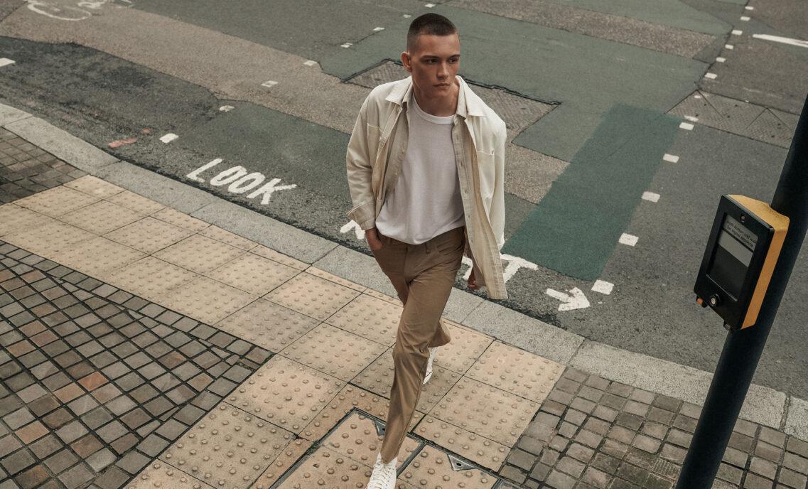 A person walking on the sidewalk looking over their shoulder. They have a hand in their pocket and are wearing a beige Dockers® button up over a white T-shirt and tan Dockers® GO pants.