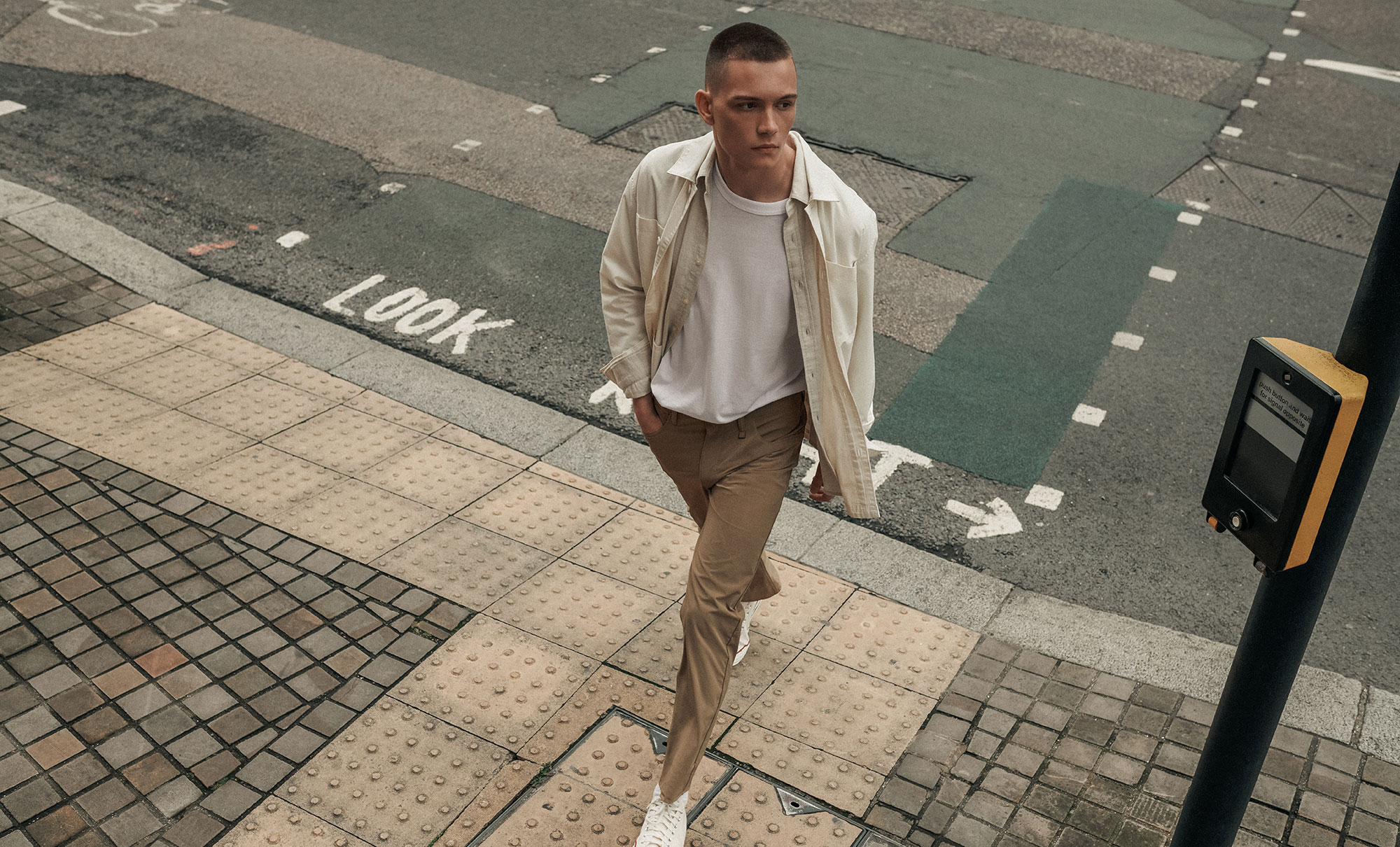 A person walking on the sidewalk looking over their shoulder. They have a hand in their pocket and are wearing a beige Dockers® button up over a white T-shirt and tan Dockers® GO pants.