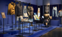 A section of the new Levi's® exhibition, Icons, Innovations & Firsts — Stories of Heritage and Progress From the Levi’s® Archives. Various Levi's® pieces are displayed in a dark room with a blue background. Description plaques are placed in front of each item.
