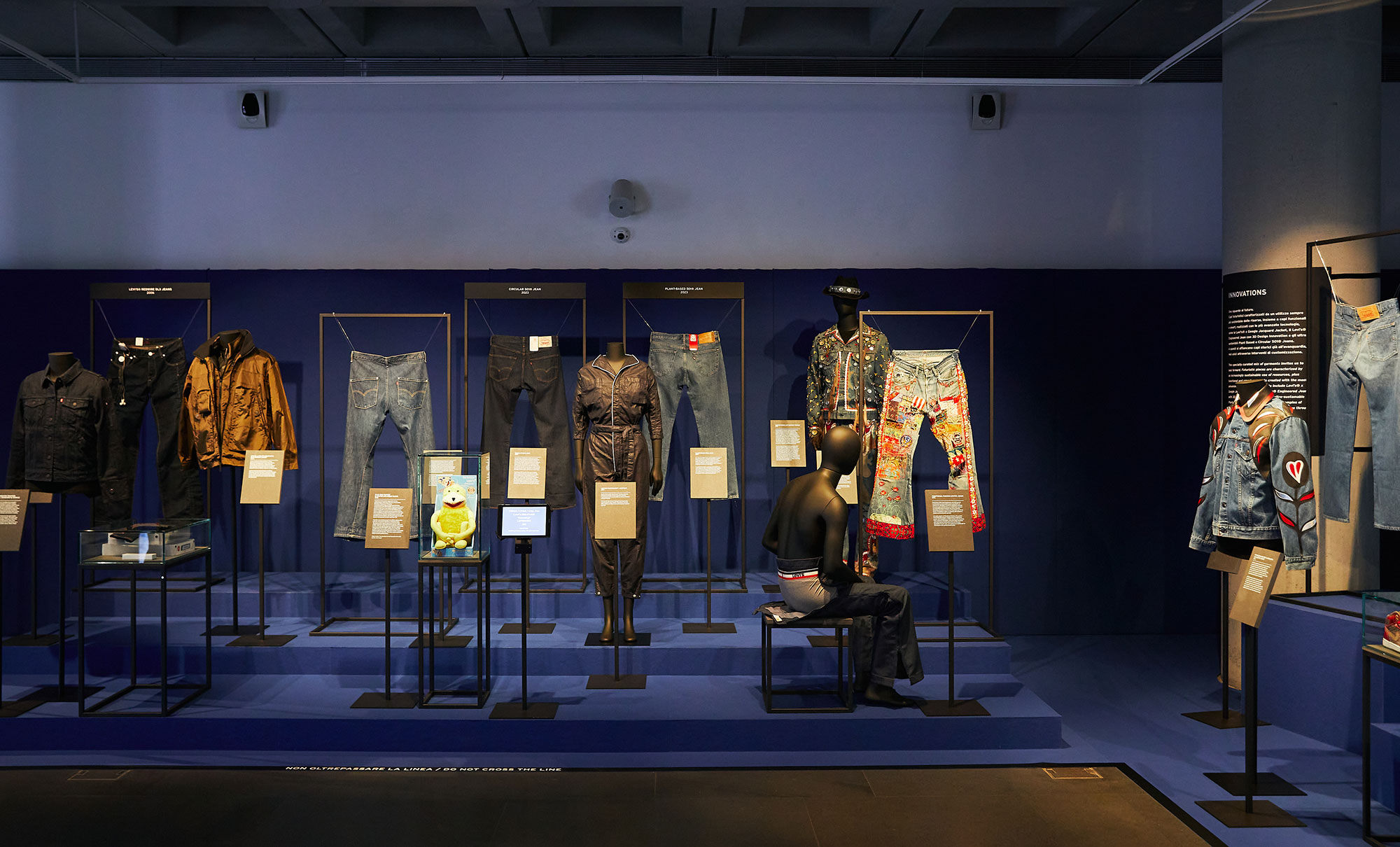 A section of the new Levi's® exhibition, Icons, Innovations & Firsts — Stories of Heritage and Progress From the Levi’s® Archives. Various Levi's® pieces are displayed in a dark room with a blue background. Description plaques are placed in front of each item.