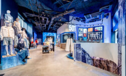 The interior of the Levi's® Champs-Élysées store in Paris, featuring mannequins dressed in head-to-toe Levi's® products, a Levi's® Tailor Shop, exposed ceiling and a 501® sign on the wall. A staircase on the right leads downstairs. 