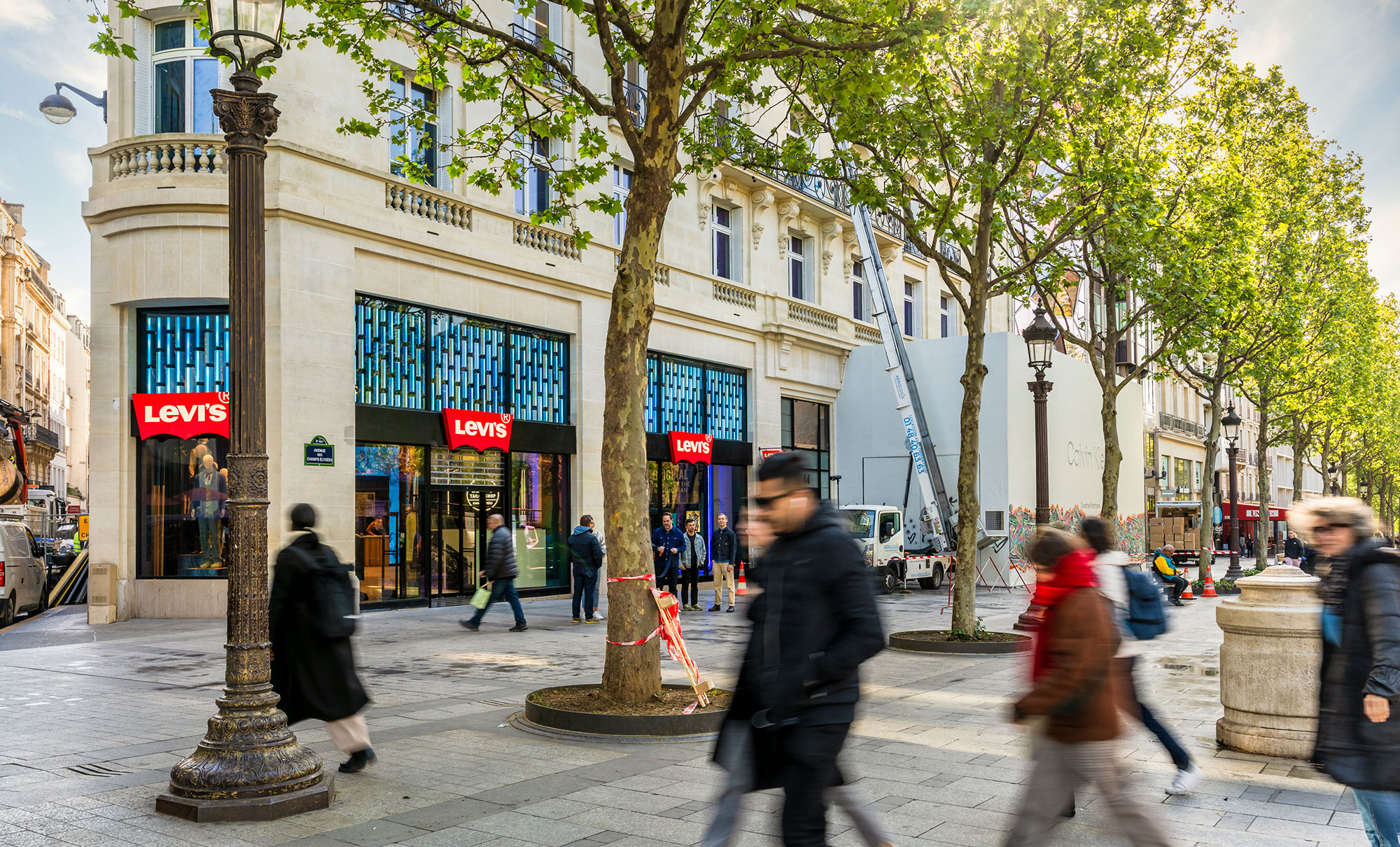 The exterior of the new Levi's® flagship store in Paris, Levi's® Champs-Élysées. A cream colored Parisian style building with large windows and the red Levi's® batwing logo placed throughout. Trees line the sidewalk and people walk around.