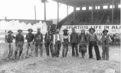 A black and white image of eleven people standing in a rodeo arena wearing Western wear. 