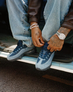 A pair of hands reaches down to tie a pair of Levi's® x Nike Air Max 95 shoes on their feet. They are wearing denim Levi's® jeans and brown long sleeve shirt. There is a wrist watch on their left wrist and a bracelet on their right wrist.