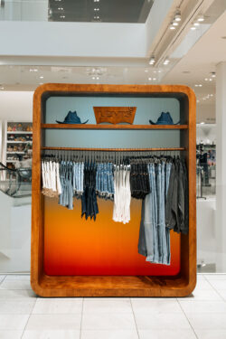 An assortment of Levi's products hang on a clothes rack within a wooden display case. A top shelf holds two denim Levi's® cowboy hats and a wooden Levi's® batwing logo.