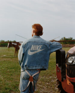 A person stands in a grass field with their back to the camera leaning against the hub of a truck. They are wearing a denim Levi's® Trucker jacket from the Levi's® x Nike collaboration featuring a Nike logo on the back.