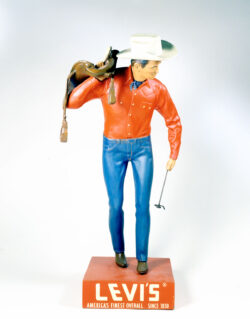 “The Saddleman,” a cowboy figure dressed in Levi’s® jeans, a Western sawtooth shirt, kerchief, cowboy hat and boots. slinging a saddle over one shoulder and holding an “LS” brand in the other like a rancher.