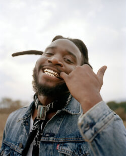 Shaboozey grins at the camera while his pinky points at a tooth grill. He wears a Levi's® Trucker jacket and bandana around his neck.