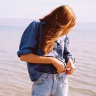 A person stands in front of an ocean with their head down and hair covering their face. They are wearing a long sleeve button down blue top with the sleeves rolled up, tucked into light wash blue denim jeans. Their hands are at their waistband.