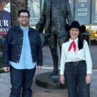 Levi's store manager Jesse Sandhu and LS&Co. historian Tracey Panek smile and stand in front of the Buffalo Bill Center of the West in Cody, Wyoming. A poster in the back promoting the featured exhibition reads "Spur of the moment: Iconic styles of the American West." Jesse wears a dark was Levi's® denim Trucker jacket over a blue button up shirt and dark was Levi's® jeans. Tracey wears a black cowboy hat, white button up shirt, red ascot scarf and Levi's® jeans.