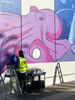 Antoine Marnata of 1AM Projects stands by a cart of paint and paints a purple octopus on a section of a mural
