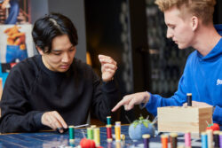 A person wearing a black hoodie sits at a blue-tiled table covered with spools of thread. They hold a needle and thread in one hand and look down at a fabric swatch. A person to the right in a blue hoodie points down at the swatch.