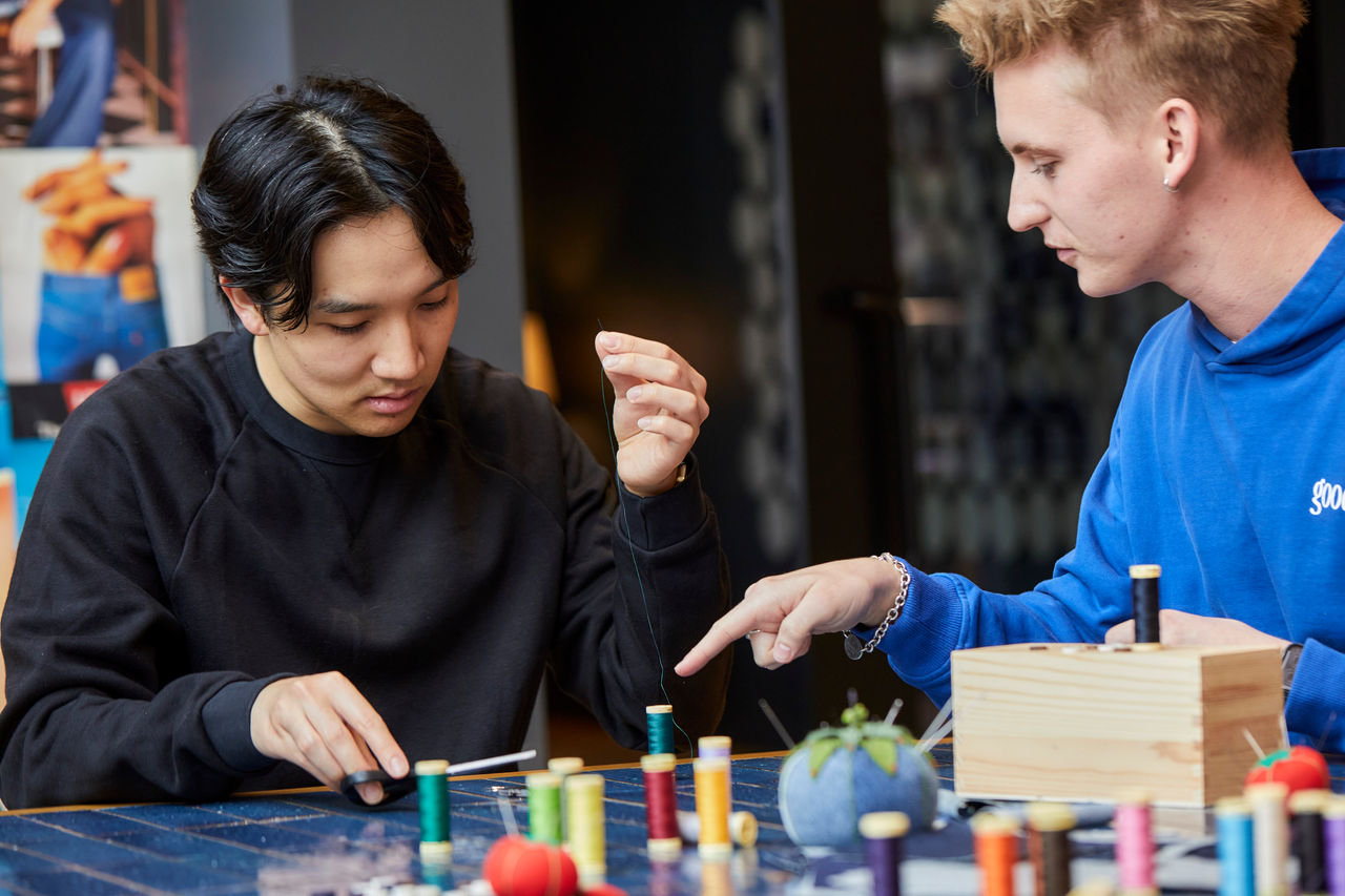 A person wearing a black hoodie sits at a blue-tiled table covered with spools of thread. They hold a needle and thread in one hand and look down at a fabric swatch. A person to the right in a blue hoodie points down at the swatch.