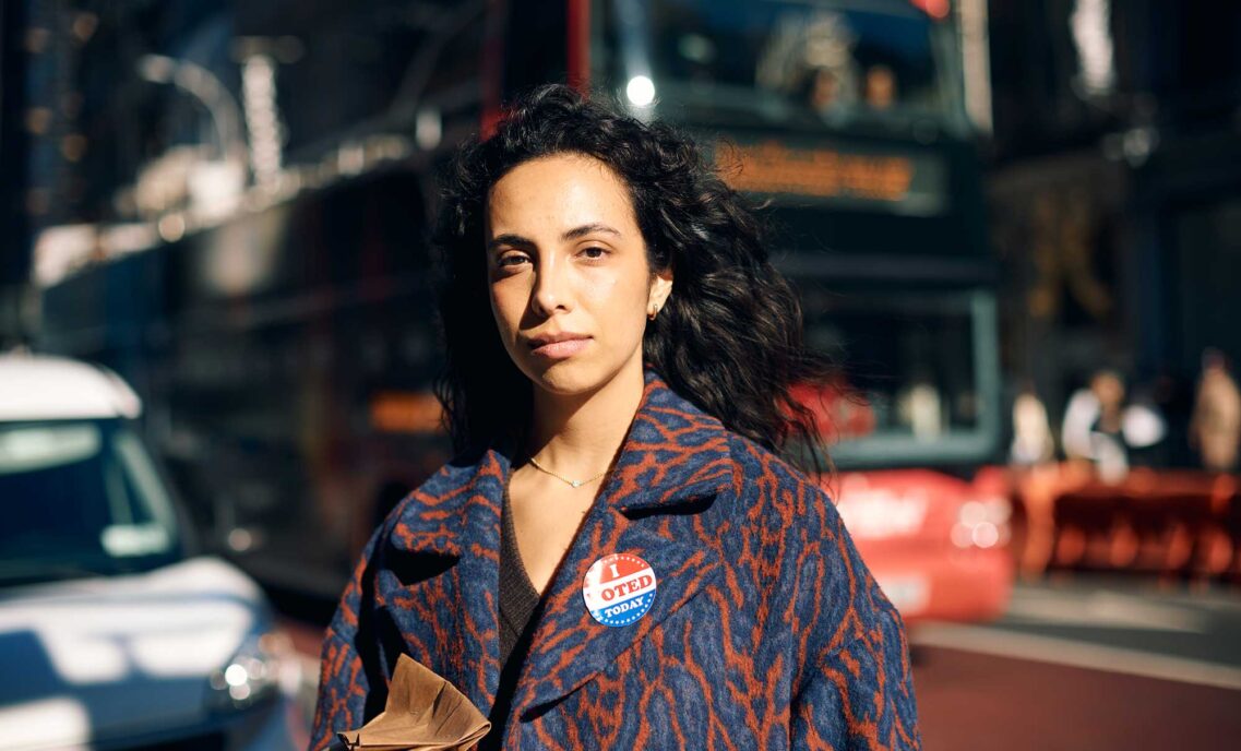 A person wearing an abstract blue and red patterned jacket and an "I Voted Today" sticker looks into the camera in front of a blurred-out urban city background.