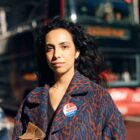 A person wearing an abstract blue and red patterned jacket and an "I Voted Today" sticker looks into the camera in front of a blurred-out urban city background.
