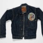 A WWII-era Levi's® denim jacket photographed on top of a plain grey background. The jacket features then name "Betty Jane Moore" written on the inside collar patch above the text "Fairfield Suisun Army Air Base," along with laurel leaf donut buttons, a single front pocket without a flap, a 13th Air Transport Squadron patch sewn on the front, and visible wear patterns, including the indigo dye rubbed off at the collar.