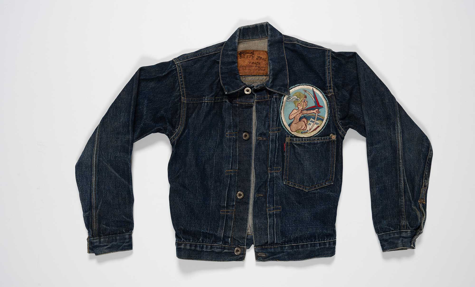 A WWII-era Levi's® denim jacket photographed on top of a plain grey background. The jacket features then name "Betty Jane Moore" written on the inside collar patch above the text "Fairfield Suisun Army Air Base," along with laurel leaf donut buttons, a single front pocket without a flap, a 13th Air Transport Squadron patch sewn on the front, and visible wear patterns, including the indigo dye rubbed off at the collar.