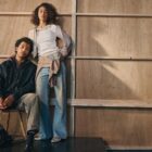 A person poses in a chair wearing a dark Levi's® jacket and Levi's® jeans. Another person standing up leans on the person in a chair. They wear a Levi's® top, Levi's® jeans and a Levi's® sweatshirt tied around their waist. The two people pose in front of a wooden wall.