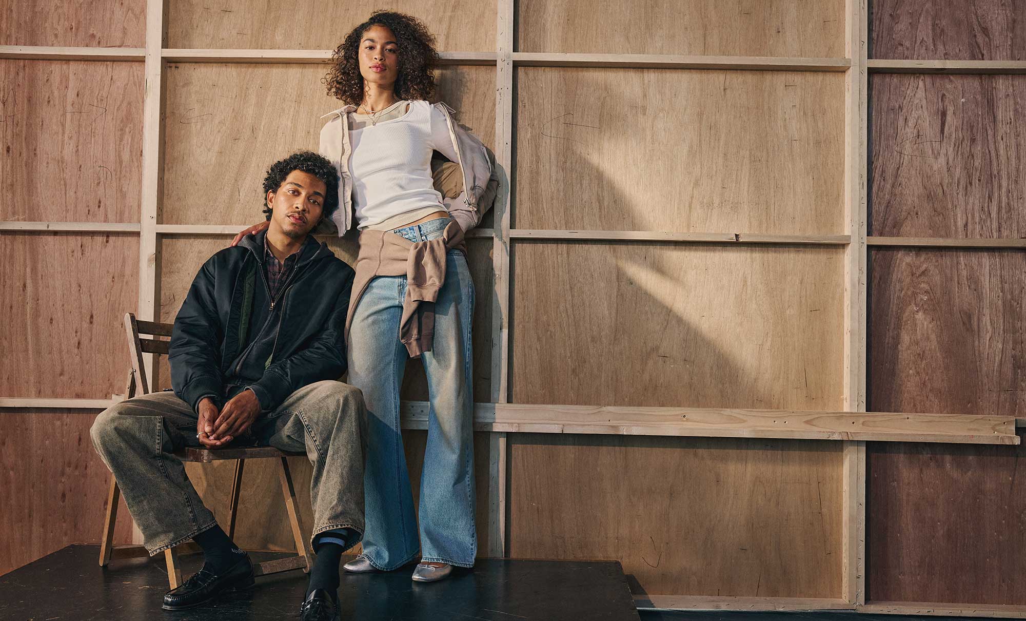 A person poses in a chair wearing a dark Levi's® jacket and Levi's® jeans. Another person standing up leans on the person in a chair. They wear a Levi's® top, Levi's® jeans and a Levi's® sweatshirt tied around their waist. The two people pose in front of a wooden wall.