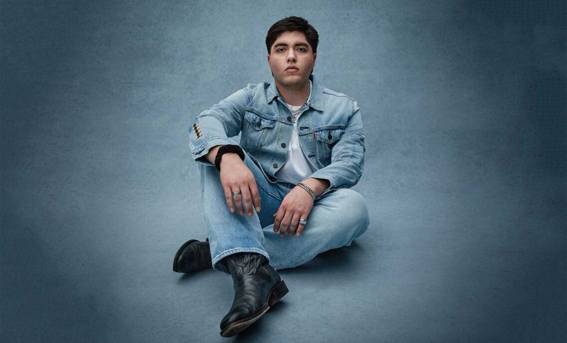 Folk singer‑songwriter Kevin Kaarl poses in a Levi's® denim jacket, Levi's® jeans and black boots against a blue background for the Levi’s® and COLORSxSTUDIOS global music program, EMERGENT.